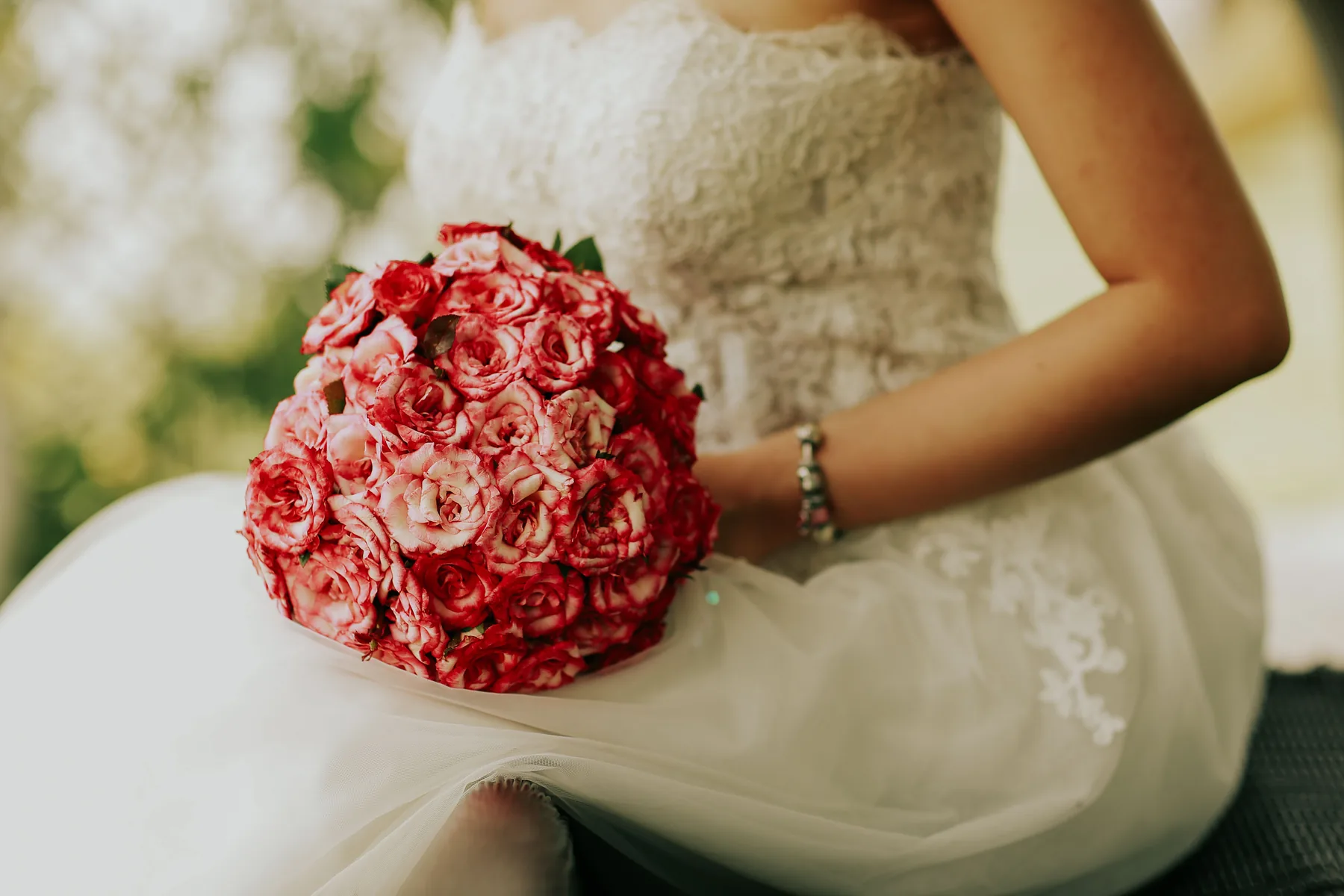 A bride seated in her dress, holding a bouquet of red and pink roses