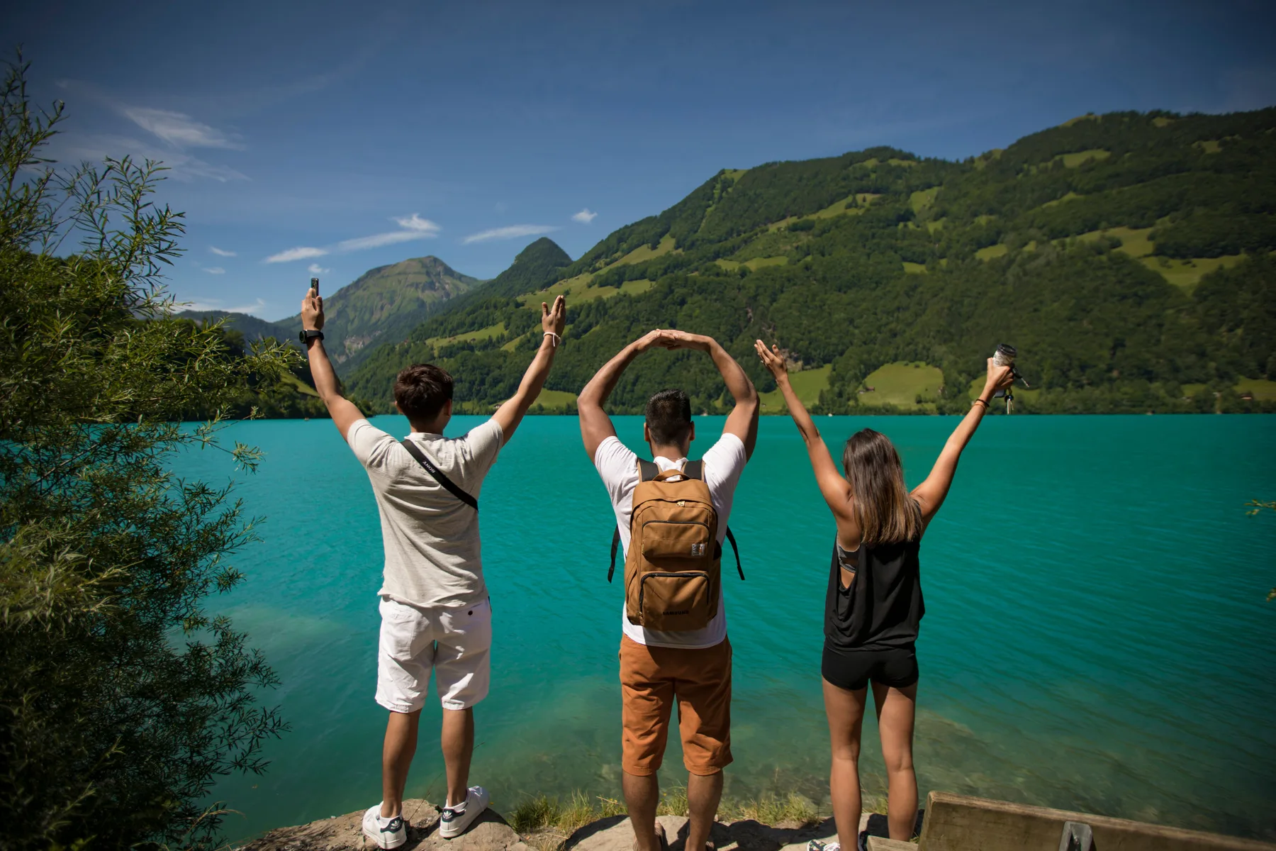Three friends with their arms raised, facing a turquoise mountain lake