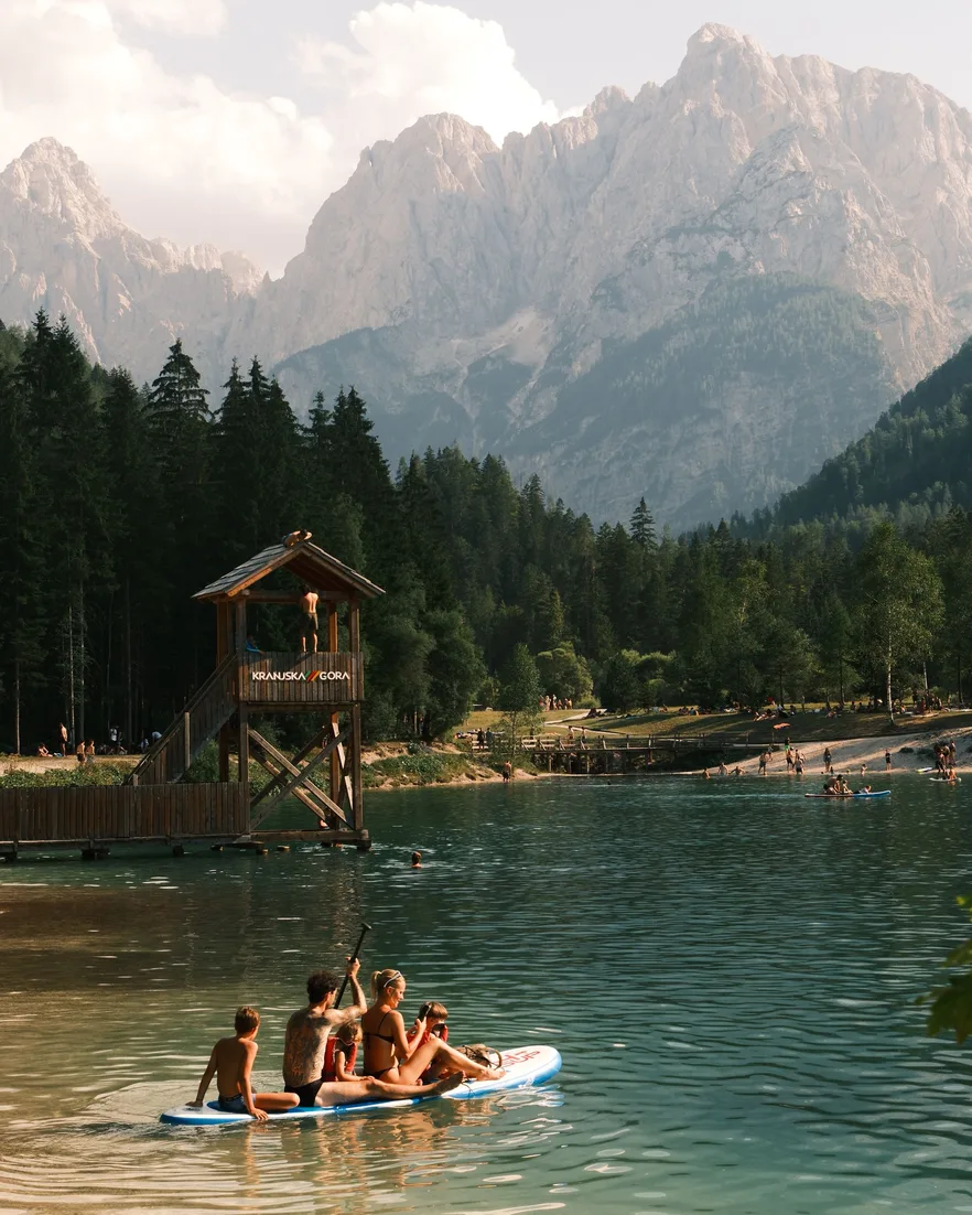Four friends sharing a paddleboard on a green alpine lake below rocky peaks