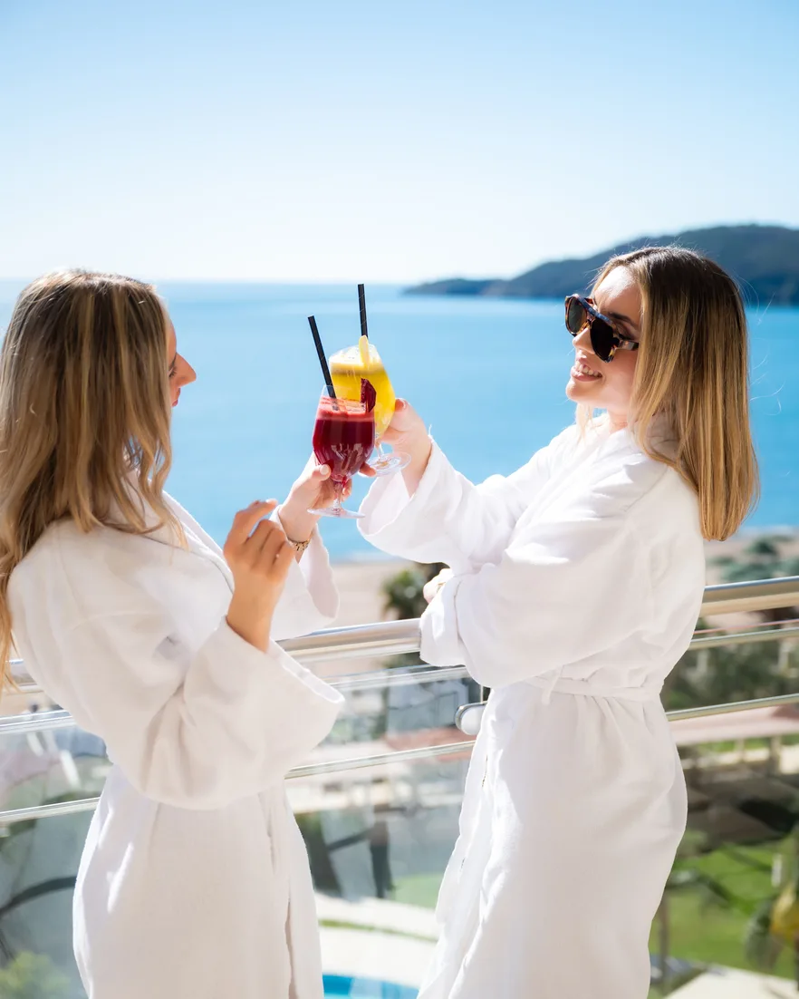 Two friends in white robes toasting with cocktails on a hotel balcony above the sea