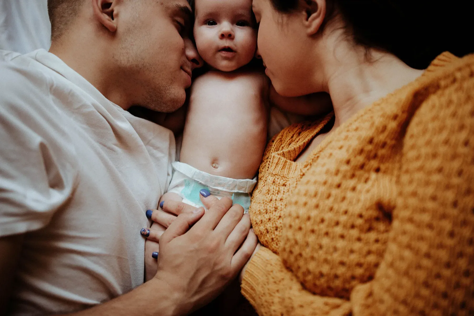 Two parents lying either side of their baby, each kissing the baby's cheek