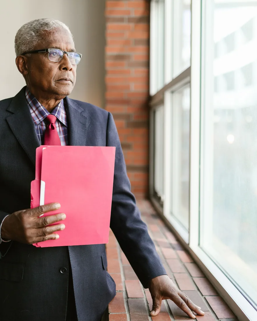 An older man in a suit holding a folder, looking out of an office window